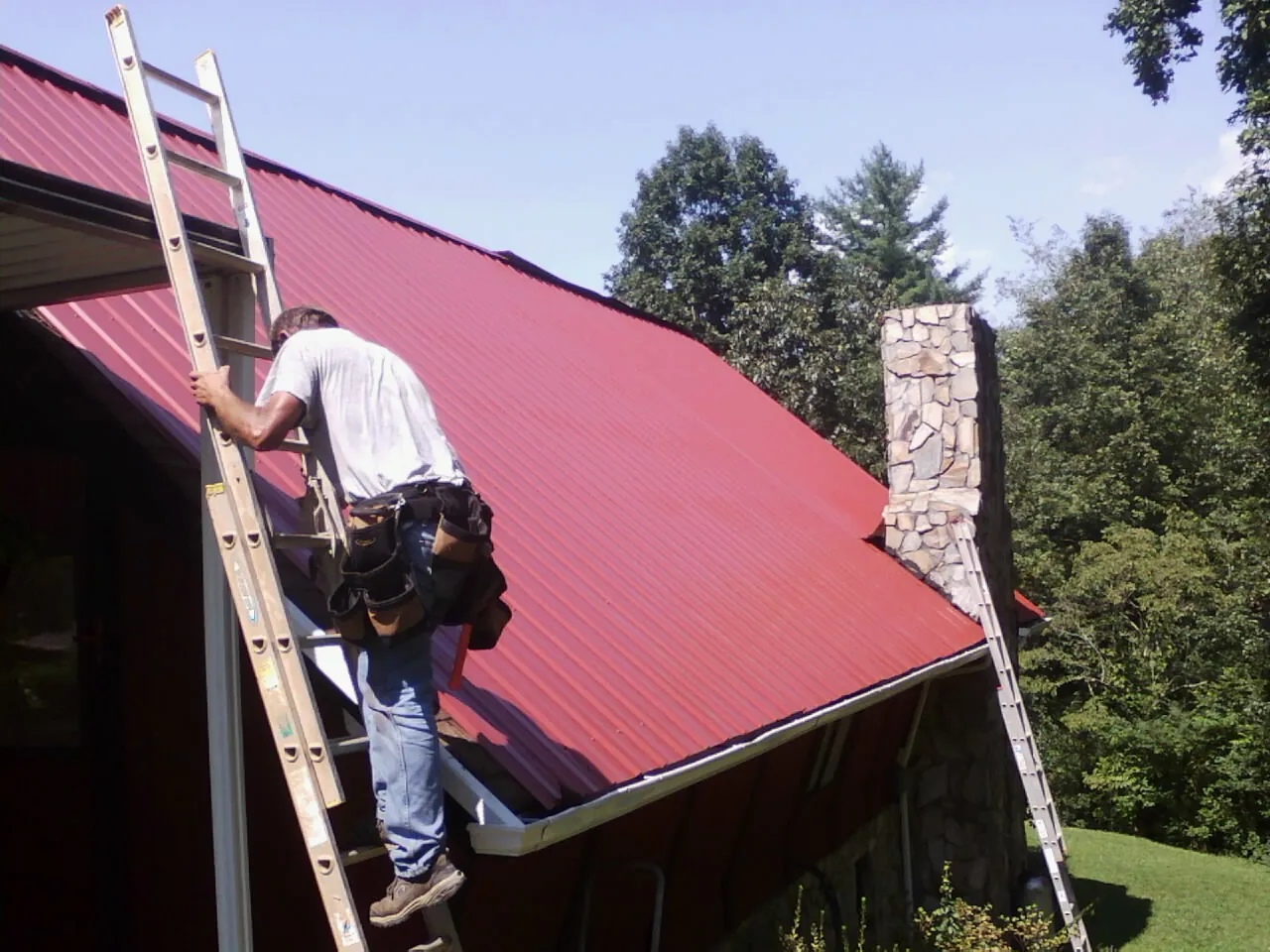 Roofer on a ladder beside a red metal roof and stone chimney — Soffit & Fascia Repair by Roofing Brothers in Cedarburg